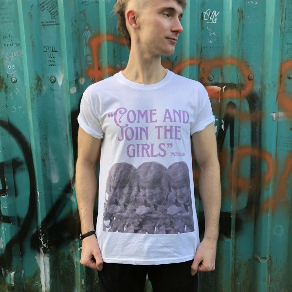 A male model wearing the 'Come And Join The Girls' T-shirt with a green metal background behind him. The background has various graffiti.